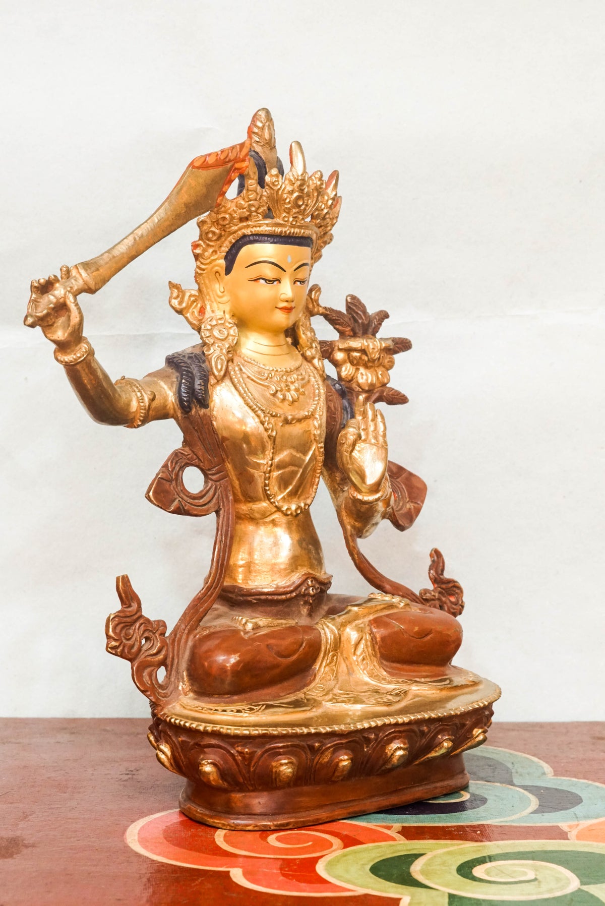 Gilt bronze statue of Manjushri, the Bodhisattva of Wisdom, seated in vajra posture on a lotus base, raising the flaming sword of wisdom in his right hand and holding a lotus stem supporting the Prajñāpāramitā scripture in his left, crafted in traditional Himalayan Buddhist style.