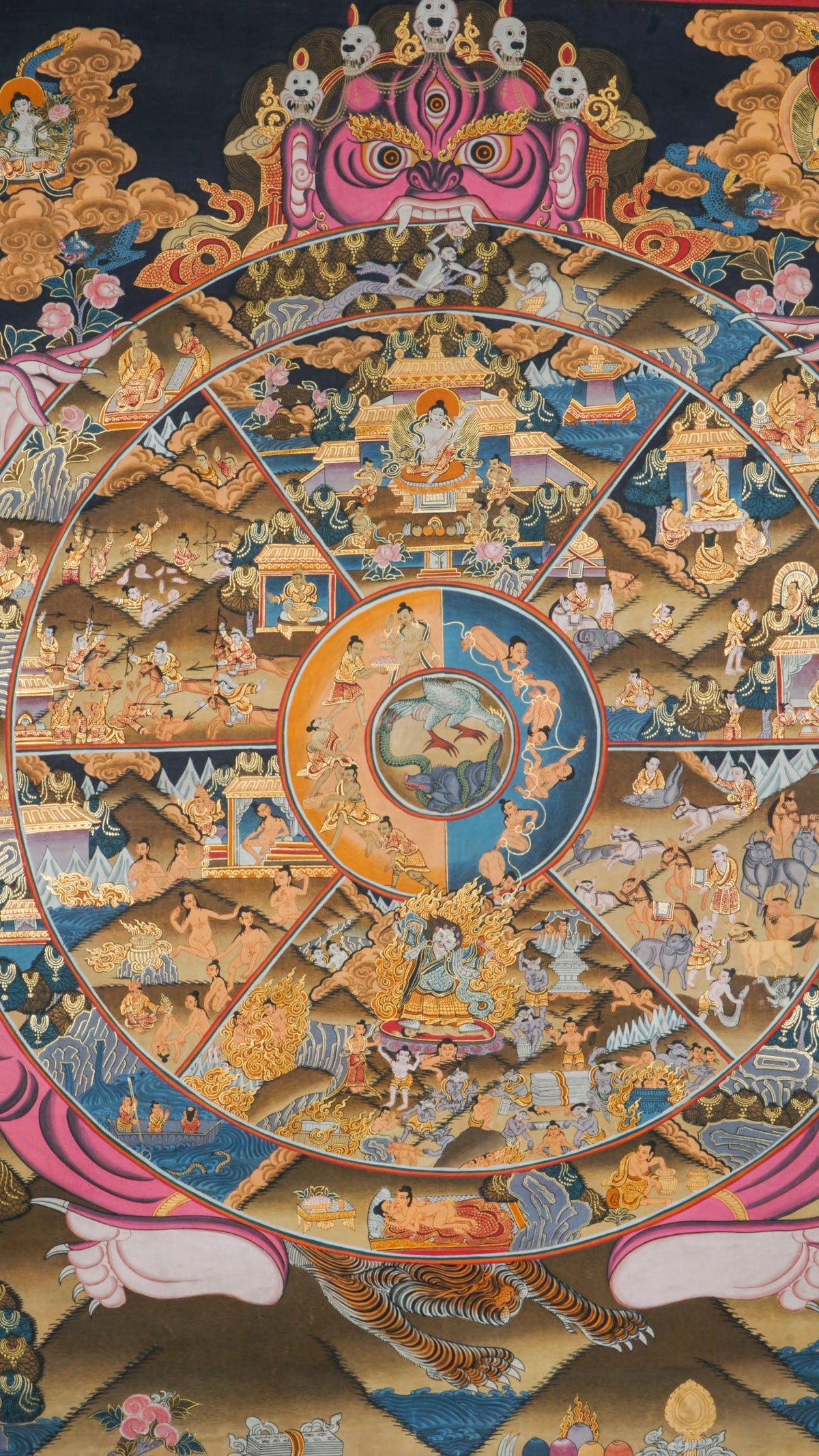 Detailed thangka-style painting showing a large circular wheel divided into multiple wedge-shaped sections filled with small human figures and scenes, surrounded by concentric rings, held by a large multi-eyed figure at the top, with clouds, flames, and ornate decorative borders in dark blue, gold, pink, and brown tones.