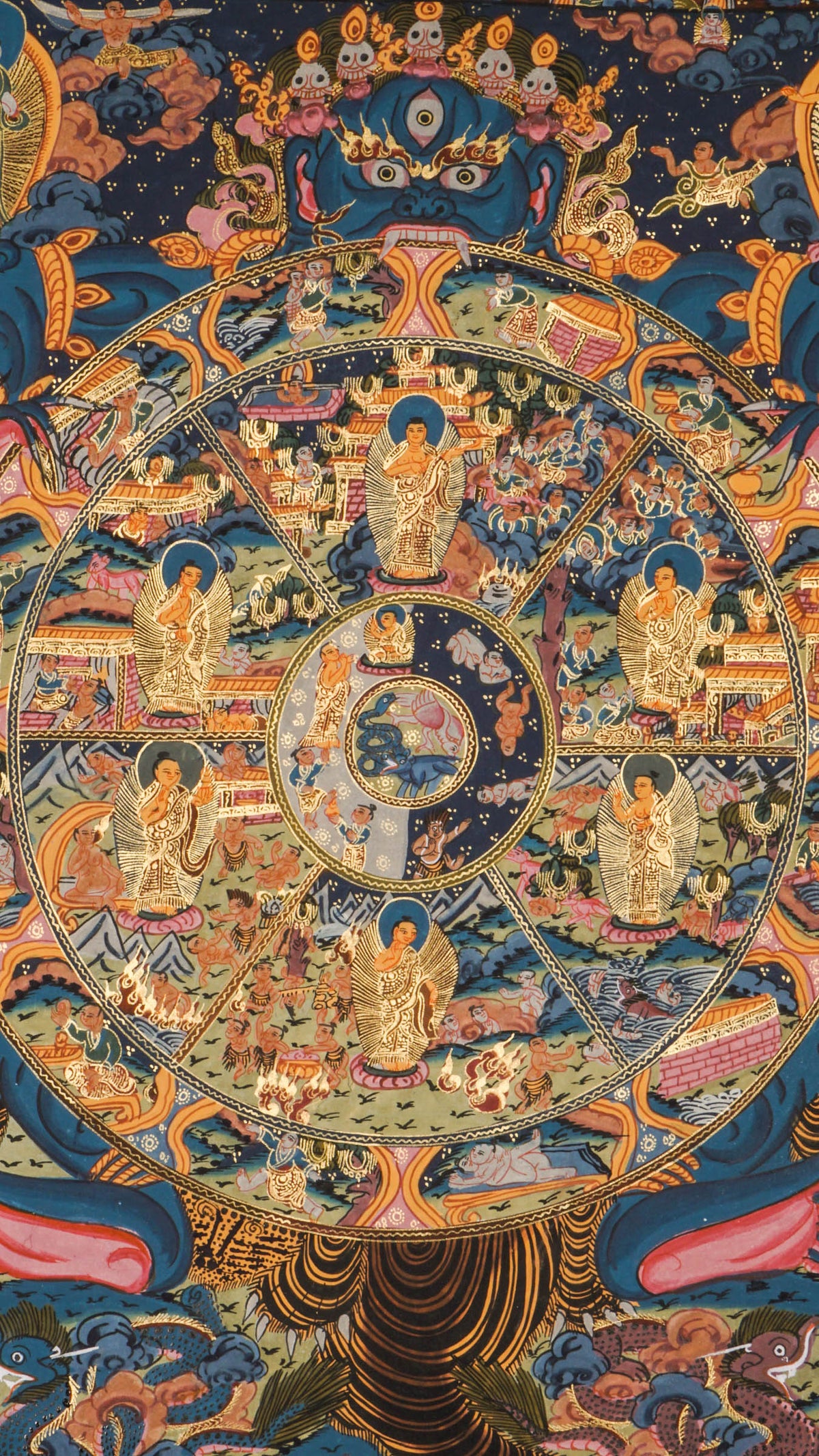 Detailed thangka-style painting showing a large circular wheel divided into multiple sections filled with small human figures, animals, buildings, and landscape scenes, surrounded by concentric decorative rings and held by a large blue multi-eyed figure with extended arms, set against a dark background with clouds, flames, and ornate gold, red, and blue patterns.
