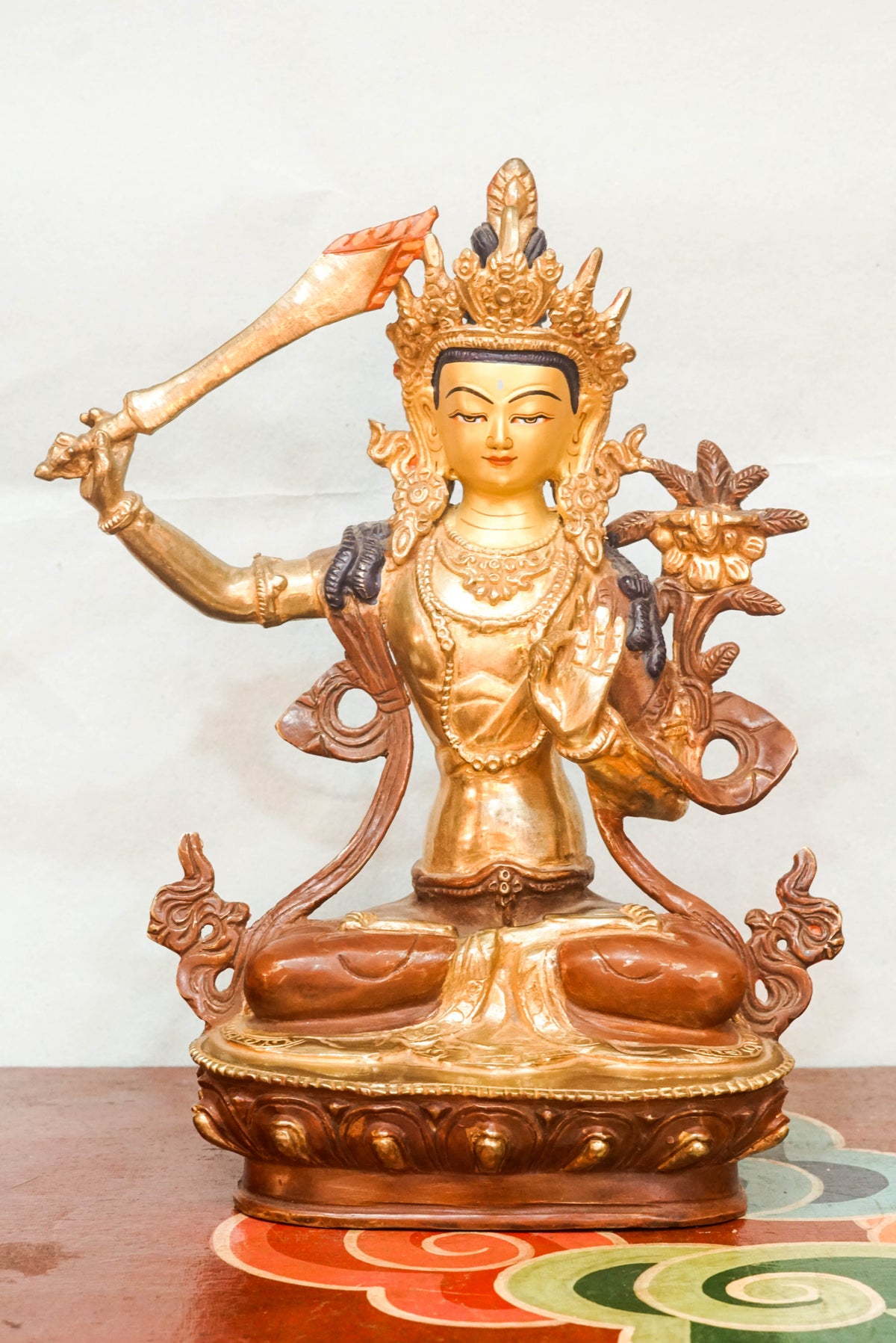 Gilt bronze statue of Manjushri, the Bodhisattva of Wisdom, seated in vajra posture on a lotus base, raising the flaming sword of wisdom in his right hand and holding a lotus stem supporting the Prajñāpāramitā scripture in his left, crafted in traditional Himalayan Buddhist style.