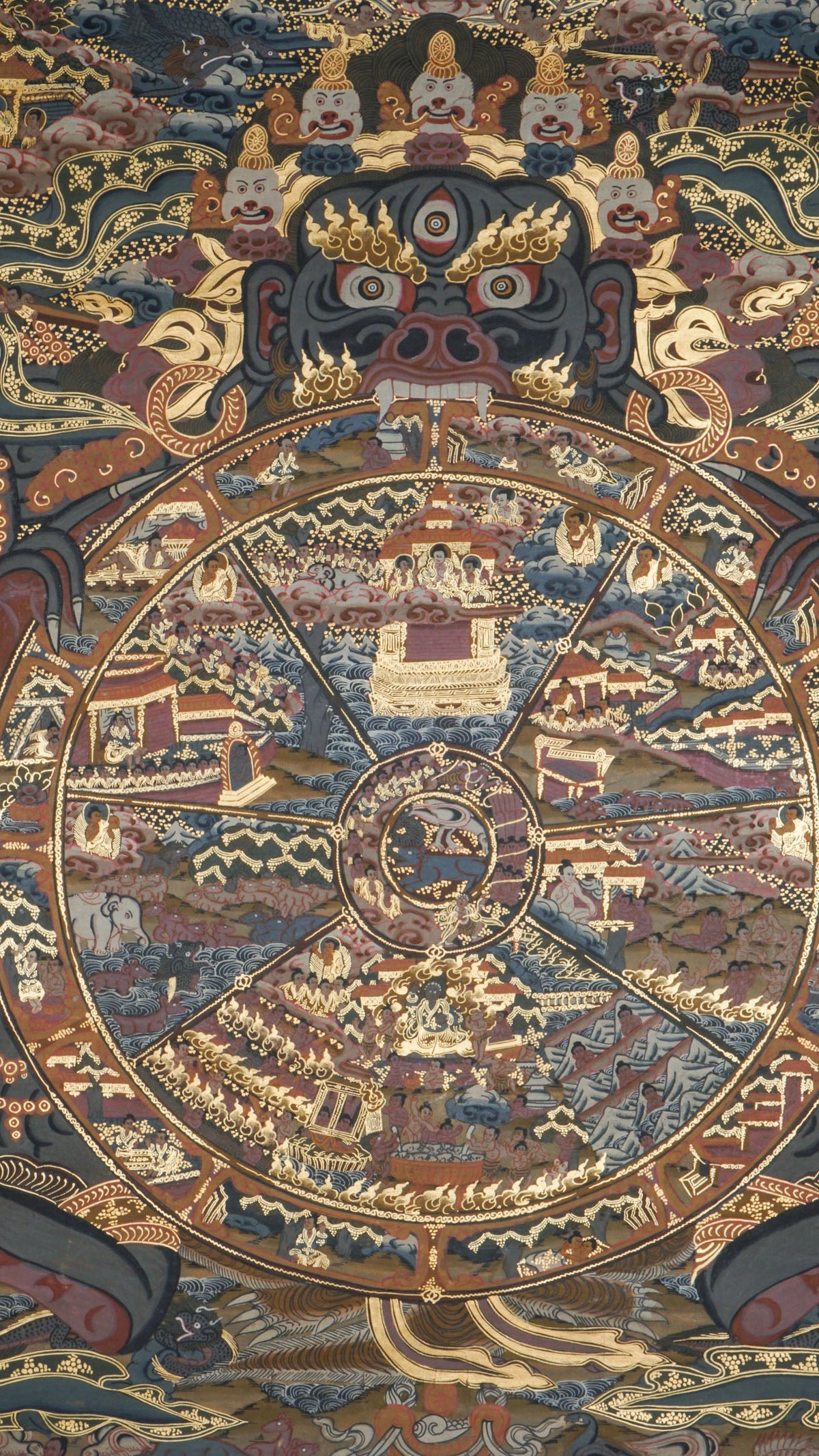 Detailed thangka-style painting showing a large circular wheel divided into multiple wedge-shaped sections filled with small human figures, buildings, animals, and landscape scenes, surrounded by concentric decorative rings. The wheel is held by a large dark blue multi-eyed figure with extended arms and clawed hands, set against a richly patterned background with clouds, flames, and ornate gold, brown, red, and blue detailing.